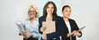 © Prostock-studio - Three businesswomen stand together in a modern office space. Each holds important documents and displays a confident smile, showcasing professionalism and teamwork in their environment.
