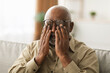 © Prostock-studio - An older man sits on a couch, rubbing his eyes while wearing glasses. The room is bright and cozy, indicating a moment of rest and reflection in his day.