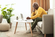 © Prostock-studio - A thoughtful elderly man is engaged in studying while comfortably seated on a sofa in a bright living room. He writes notes in a notebook with a laptop in front of him.