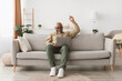 © Prostock-studio - An elderly man is sitting on a light gray couch, engaged with his laptop. He appears focused while enjoying leisure time in a bright, comfortable living room decorated with plants and warm light.