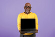 © Prostock-studio - An older man stands with a smile while holding a laptop in front of him. He wears a yellow sweater and looks directly at the camera against a purple background.