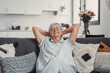 © Daniel - Elderly woman relaxes on a sofa at home, stretching and smiling peacefully. She enjoys a calm and carefree moment, embracing happiness and serenity in her domestic lifestyle.