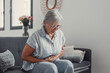 © Daniel - Elderly woman sits on a sofa at home holding her stomach, experiencing abdominal pain and discomfort. She looks tired and stressed while managing digestive issues during her daily life.