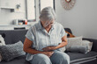 © Daniel - Elderly woman sits on a sofa at home holding her stomach, experiencing abdominal pain and discomfort. She looks tired and stressed while managing digestive issues during her daily life.