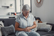 © Daniel - Elderly woman sits on a sofa at home holding her stomach, experiencing abdominal pain and discomfort. She looks tired and stressed while managing digestive issues during her daily life.