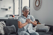 © Daniel - Elderly woman sits on a sofa at home holding her chest in discomfort. She appears worried and fatigued, showing signs of heart pain and anxiety during a stressful moment.