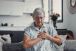 © Daniel - Elderly woman sits on a sofa at home holding her chest in discomfort. She appears worried and fatigued, showing signs of heart pain and anxiety during a stressful moment.