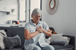 © Daniel - Elderly woman sits on a sofa at home holding her chest in discomfort. She appears worried and fatigued, showing signs of heart pain and anxiety during a stressful moment.