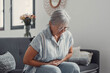 © Daniel - Elderly woman sits on a sofa at home holding her stomach, experiencing abdominal pain and discomfort. She looks tired and stressed while managing digestive issues during her daily life.