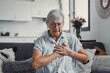 © Daniel - Elderly woman sits on a sofa at home holding her chest in discomfort. She appears worried and fatigued, showing signs of heart pain and anxiety during a stressful moment.