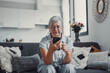 © Daniel - Elderly woman sits on a sofa at home using her smartphone to browse and shop online. She focuses on digital communication and enjoys a modern e-commerce lifestyle.