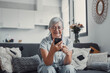 © Daniel - Elderly woman sits on a sofa at home using her smartphone to browse and shop online. She focuses on digital communication and enjoys a modern e-commerce lifestyle.