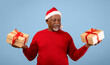 © Prostock-studio - A man wearing a red sweater and a Santa hat stands holding two gift boxes. He appears to be celebrating a festive occasion. The background is light blue and accentuates the gifts he is holding.