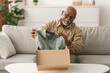 © Prostock-studio - An elderly man is smiling while unpacking a freshly delivered shirt from a cardboard box in his cozy living room. Sunlight fills the space, creating a warm atmosphere.