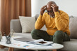 © Prostock-studio - Man with gray hair sits on a couch, looking troubled as he rubs his temples. Papers and a laptop are on the table, showcasing signs of a challenging workday at home.