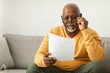 © Prostock-studio - A mature man with gray hair sits on a light gray couch, thoughtfully reading a piece of paper. He wears a yellow sweater and looks focused, adjusting his glasses as he engages with the content.