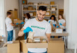 © Prostock-studio - A millennial male volunteer stands proudly with a food donation box filled with grains, beans, and canned food. He is in a charity center, smiling and happy to help as others pack donations nearby.