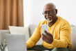 © Prostock-studio - An older man is engaged in a digital activity, holding a smartphone while seated on a comfortable couch. A laptop rests in front of him in a warm, inviting living room.