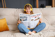 © Pixel-Shot - Young woman in headphones reading newspaper on sofa at home