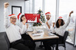 © sofiko14 - Team of multiethnic business people in Santa hats raising hands and screaming with happiness looking at camera while working together in decorated with Christmas tree office in business center.