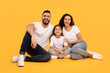 © Prostock-studio - A cheerful family sits on the floor against a vibrant yellow background. The man, woman, and young girl are dressed in casual clothing while sharing joyful expressions and laughter.