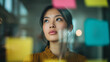 © INSPIRIA.K - A young Asian focused woman looking up through a glass wall covered in colorful sticky notes, symbolizing deep thought, creative planning, and business strategy