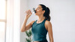© Umairey - Fit young woman in athletic wear pauses her workout to drink refreshing water from a clear bottle indoors with bright natural light streaming through a window