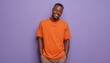 © Tetiana - Smiling Young African American Man In 20s Wearing Orange T-Shirt, Holding Hands In Pockets, And Looking At Camera In Pastel Violet Studio Portrait.