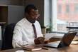 © DragonImages - Middle aged Black man sitting at desk reading document, wearing glasses and formal attire, working in office with laptop and notebook, school principal reviewing paperwork