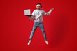 © Prostock-studio - A young person wearing a Christmas hat jumps in excitement while holding a laptop. The vibrant red background enhances the festive mood, showcasing joy and celebration.
