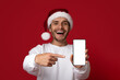© Prostock-studio - A man wearing a Santa hat smiles widely while pointing at his smartphone with a blank screen. The bright red background adds a festive touch to the scene.