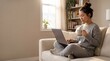 © Николай Срибяник - Focused young Asian woman sitting cross legged on sofa using laptop computer and holding coffee cup in sunny cozy living room while working from home