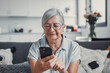 © Daniel - Elderly woman sits on a sofa at home using her smartphone to browse and shop online. She focuses on digital communication and enjoys a modern e-commerce lifestyle.