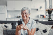 © Daniel - Elderly woman smiles while sitting on a sofa at home using her smartphone for online shopping. She enjoys a cheerful and connected lifestyle while exploring digital marketplaces.