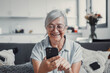 © Daniel - Elderly woman smiles while sitting on a sofa at home using her smartphone for online shopping. She enjoys a cheerful and connected lifestyle while exploring digital marketplaces.