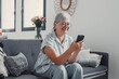© Daniel - Elderly woman smiles while sitting on a sofa at home using her smartphone for online shopping. She enjoys a cheerful and connected lifestyle while exploring digital marketplaces.