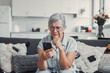 © Daniel - Elderly woman sits on a sofa at home looking at her phone with disappointment. She receives bad news while shopping online, feeling frustrated and cautious about digital transactions.