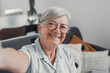 © Daniel - Elderly woman takes a selfie on a sofa at home, smiling and feeling cheerful. She enjoys a calm and peaceful moment while sharing her life on social media in a reflective way.