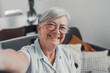 © Daniel - Elderly woman takes a selfie on a sofa at home, smiling and feeling cheerful. She enjoys a calm and peaceful moment while sharing her life on social media in a reflective way.