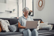 © Daniel - Elderly woman sits on a sofa at home using her computer to browse and shop online. She focuses on digital communication and enjoys a productive e-commerce routine.