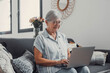 © Daniel - Elderly woman sits on a sofa at home using her computer to browse and shop online. She focuses on digital communication and enjoys a productive e-commerce routine.