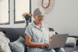 © Daniel - Elderly woman sits on a sofa at home using her computer to browse and shop online. She focuses on digital communication and enjoys a productive e-commerce routine.