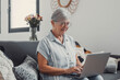 © Daniel - Elderly woman sits on a sofa at home using her computer to browse and shop online. She focuses on digital communication and enjoys a productive e-commerce routine.