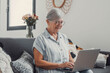 © Daniel - Elderly woman sits on a sofa at home using her computer to browse and shop online. She focuses on digital communication and enjoys a productive e-commerce routine.