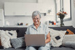 © Daniel - Elderly woman sits on a sofa at home using her computer to browse and shop online. She focuses on digital communication and enjoys a productive e-commerce routine.