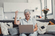 © Daniel - Elderly woman celebrates while using her computer at home after a successful online purchase. She feels cheerful and euphoric enjoying her digital victories.