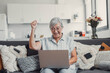© Daniel - Elderly woman celebrates while using her computer at home after a successful online purchase. She feels cheerful and euphoric enjoying her digital victories.