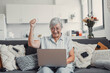 © Daniel - Elderly woman celebrates while using her computer at home after a successful online purchase. She feels cheerful and euphoric enjoying her digital victories.