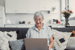 © Daniel - Elderly woman celebrates while using her computer at home after a successful online purchase. She feels cheerful and euphoric enjoying her digital victories.