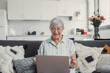 © Daniel - Elderly woman celebrates while using her computer at home after a successful online purchase. She feels cheerful and euphoric enjoying her digital victories.